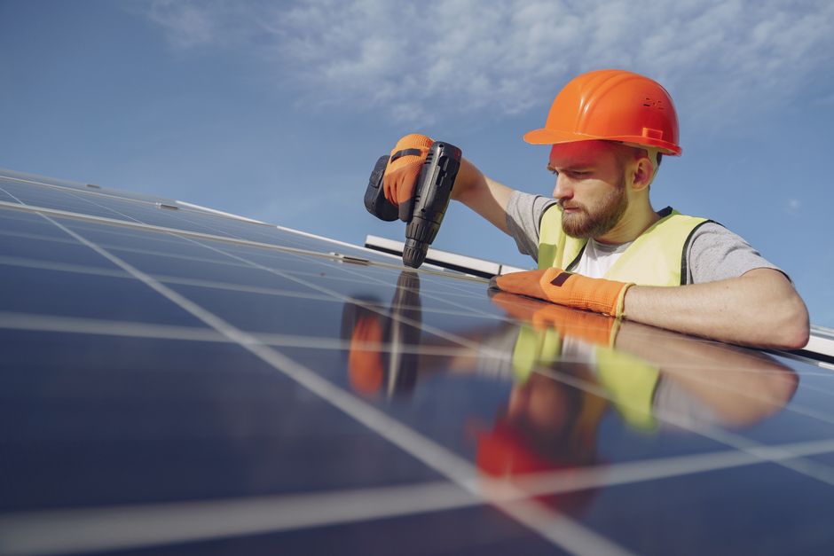 Low angle of serious bearded male electrician in hardhat and protective gloves installing solar photovoltaic panel system using a drill.