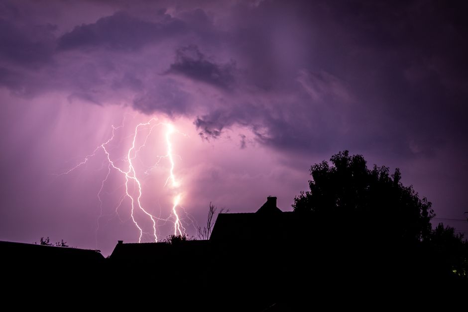 A striking lightning bolt illuminates a cloudy night sky over silhouetted houses.