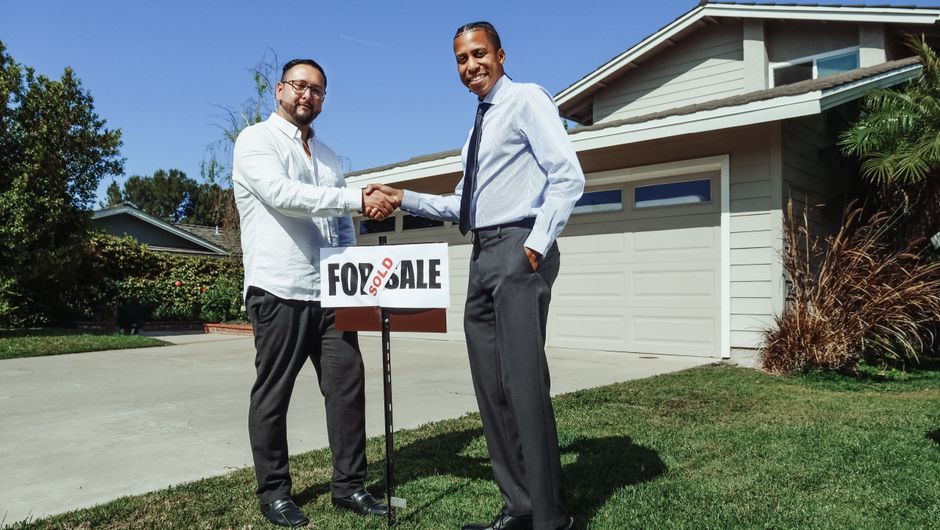 Two people shake hands in front of a new home with a sold sign, symbolizing a successful deal.