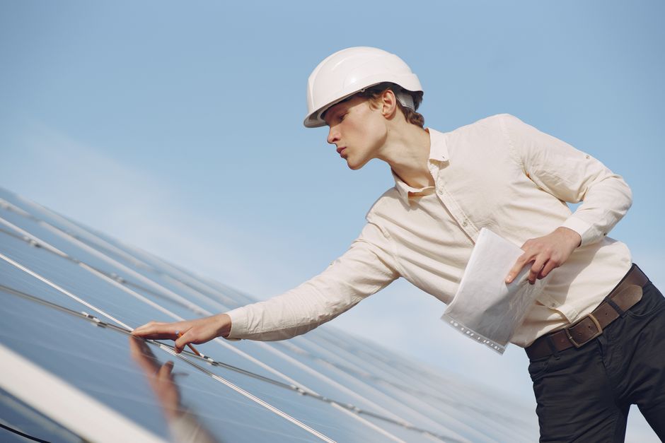 Low angle of a young inspector with documents in formal wear and hardhat standing against solar panels.